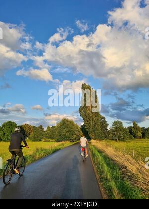 A young cyclist takes a short break on the right side of an empty road while an older cyclist passes by on the left side, recreation area of Bünde, No Stock Photo