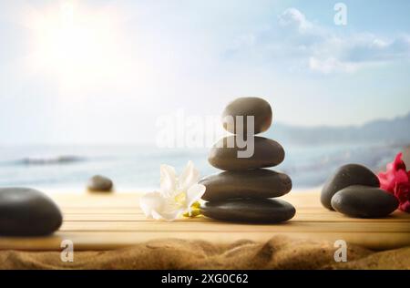 Health and body care with pile of black stones on wooden base on sand and coast background with sun. Front view. Stock Photo