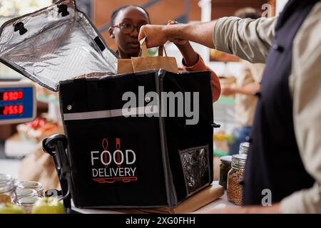 Black woman waiting for male cashier to put grocery order in thermal bag for transporting to customers. Close-up of delivery backpack being used by african american lady and storeowner at eco shop. Stock Photo