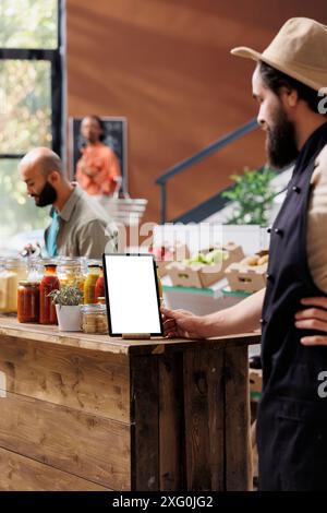 Male storekeeper wearing a hat and black apron glances at tablet with white screen placed vertically on table. Caucasian vendor using device with blank copyspace mockup template. Stock Photo
