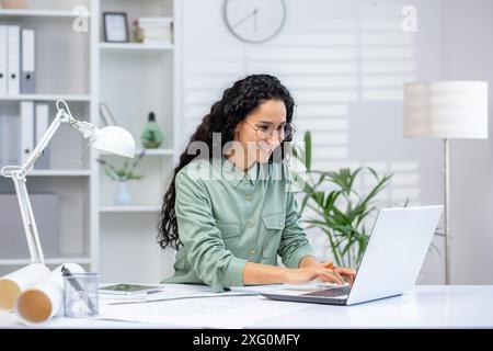 Smiling architect working on laptop in a modern, bright office filled with natural light. The workspace features stylish decor, neatly organized shelves, and indoor plants Stock Photo