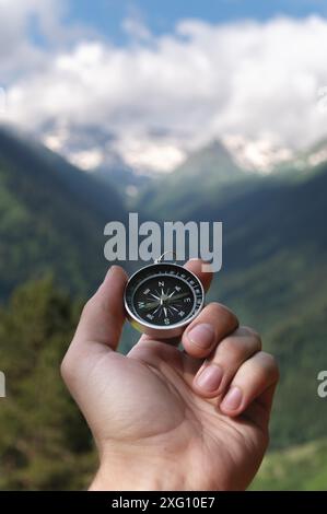 Magnetic compass in the palm of a male hand against the backdrop of a mountain range in the clouds in the summer outdoors, travel, first-person view Stock Photo