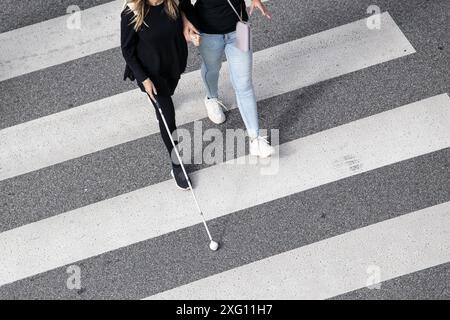 Scene of a Blind woman walking on zebra crossing helped by another person using her white cane. Help in the early stages of blindness Stock Photo