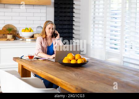 Working from home, woman using laptop and talking on smartphone in kitchen, copy space Stock Photo