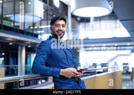 Smiling businessman holding smartphone, leaning on railing in modern office building Stock Photo