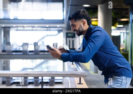 Smiling businessman using smartphone while leaning on railing in modern office, copy space Stock Photo