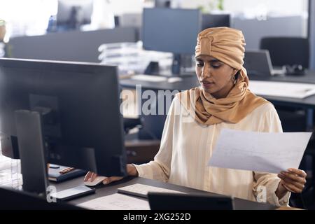 Working in office, woman reviewing documents and using desktop computer Stock Photo