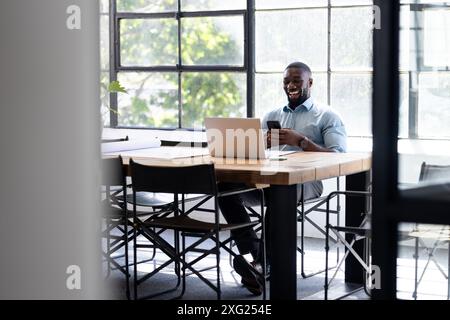 Using smartphone and laptop, man working and laughing in modern office setting Stock Photo