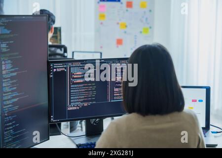 Asian software developers working on multiple screens displaying code and application diagrams in a modern office Stock Photo