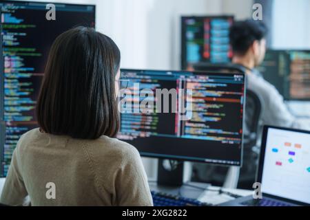 Asian software developers working on multiple screens displaying code and application diagrams in a modern office Stock Photo