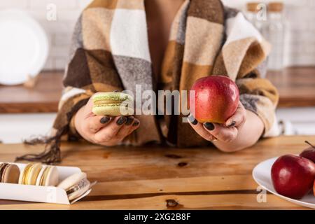 A close-up image of a womans hands holding an apple and a macaroon with a blurred background. The woman is wearing a warm scarf. The apple is red and Stock Photo