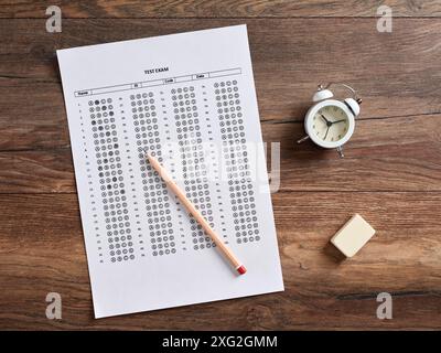 Top view of a test exam paper with alarm clock, pencil and an eraser on a wooden school desk. Education and time management in exams, quiz and school Stock Photo