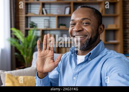 Friendly man smiling and waving hello during a video call from his cozy home setting. Ideal for concepts of virtual communication, friendliness, and remote working. Stock Photo