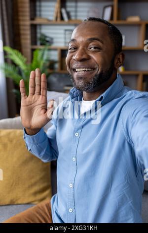 A cheerful man wearing a blue shirt waves and smiles during a video call from his living room. The warm and cozy environment suggests a relaxed and friendly conversation. Stock Photo