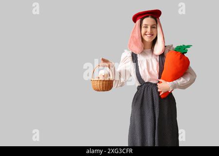 Stylish woman with bunny ears holding carrot toy and basket with Easter bread on grey background. Easter celebration Stock Photo