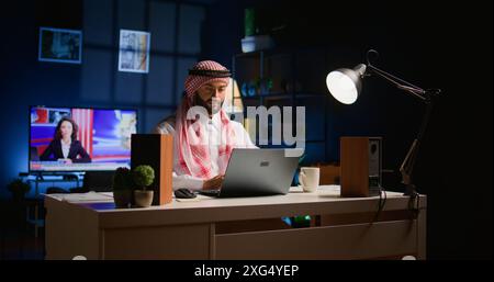 Happy Arabic employee arriving in stylish apartment personal office, starting important job tasks. Smiling Middle Eastern teleworker typing on laptop keyboard, beginning work shift at home Stock Photo