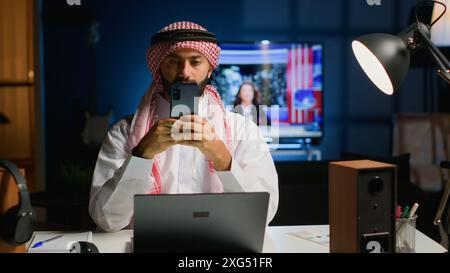 Portrait of smiling Arab freelancer at home, typing on his smartphone, surfing the net. Happy middle eastern man in traditional attire texting his friends using mobile phone Stock Photo