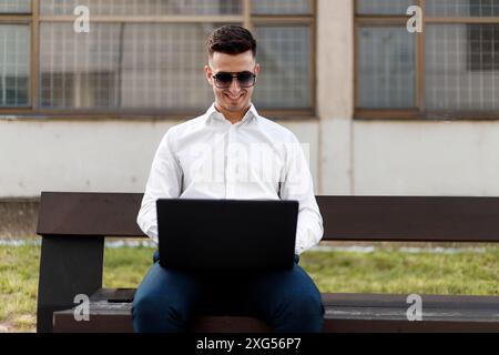 A man is sitting on a wooden bench outdoors, typing on a laptop. He is focused on the screen, with his back straight and shoulders relaxed. Stock Photo