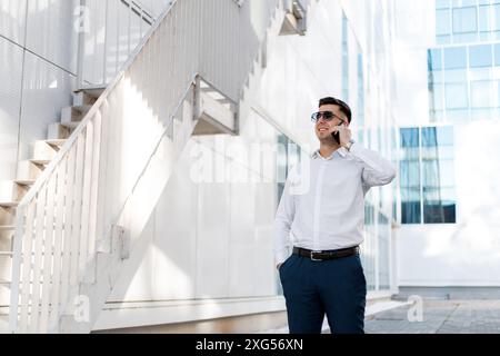A middle-aged man is standing in front of a modern office building, engaged in a phone call on his mobile device. Stock Photo