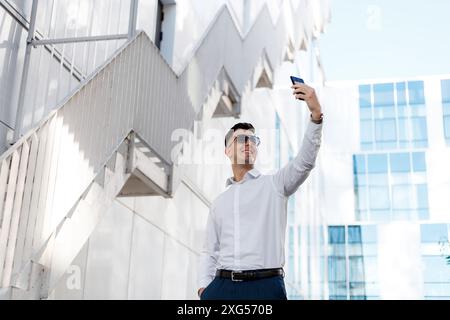 A man is seen holding up his cell phone at arms length, posing and smiling as he takes a selfie. T Stock Photo