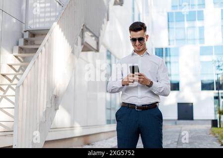 A man wearing a white shirt and blue pants is seen using a cell phone. He is standing outdoors, engrossed in the screen while tapping and swiping. Stock Photo