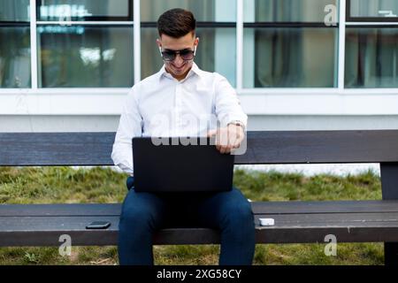 A man is sitting on a bench outdoors, engaged in using a laptop. He appears focused on the screen, typing and clicking. Stock Photo