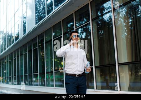 A man in a white shirt and blue pants is engaged in a phone conversation. He is standing outdoors, holding a cell phone to his ear while talking. Stock Photo