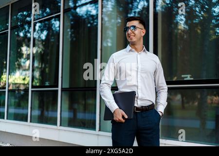 A man in casual attire standing in front of a modern office building, holding a black folder in his hand. Stock Photo