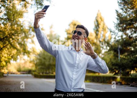 A man standing outdoors is using his cell phone to capture an image. He holds the device up, focusing on the scenery ahead as he taps the screen. Stock Photo