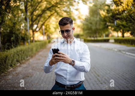 A man wearing a white shirt and blue pants is standing and staring intently at his cell phone screen. Stock Photo