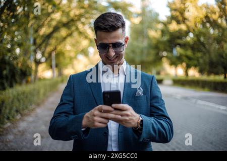 A man dressed in a suit and wearing sunglasses is focused on his cell phone screen, scrolling and typing intently. He stands in a busy urban setting. Stock Photo