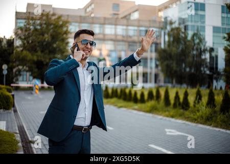 A young man in a blue suit and sunglasses stands on a city street talking on his phone with one hand while waving with the other. Stock Photo