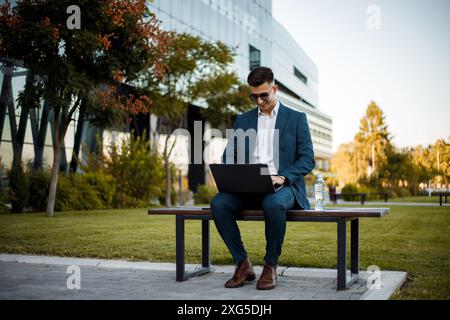 A man is sitting on a bench outdoors, focused on his laptop screen. He appears to be working or studying in a public setting. Stock Photo