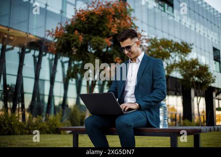 A man of Caucasian descent wearing a blue shirt and jeans sits on a wooden bench outdoors. He is focused on his laptop and typing. Stock Photo