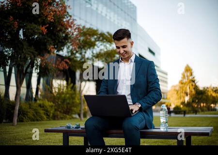 A man is sitting on a bench outdoors, focused on using a laptop. The man's hands are typing on the keyboard. Stock Photo