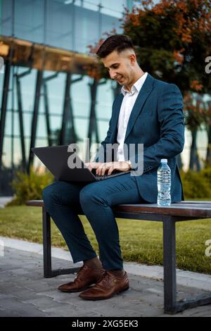 A professional man in a formal suit is seated on a bench while typing on his laptop. Stock Photo