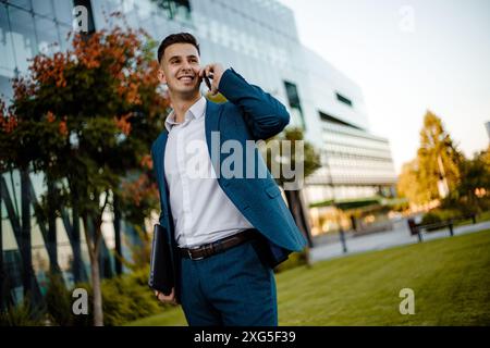 A man dressed in a formal suit is engaged in a phone conversation using a modern cell phone. He appears focused and busy, likely discussing work matte Stock Photo