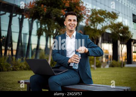 A man seated on a wooden bench outdoors, working on a laptop computer. He is focused on his screen, possibly typing or browsing. Stock Photo