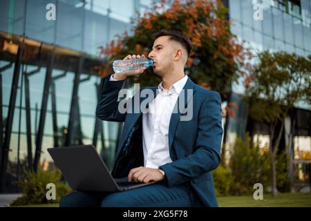 A young man in a blue suit sits outside a modern office building. He is drinking water from a plastic bottle and using a laptop. Stock Photo