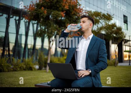 A man in a suit and white shirt sits on a bench outside a modern office building. He holds a plastic bottle of water to his mouth. Stock Photo