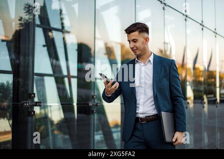 A young man in a blue suit walks down the street, looking down at his smartphone. He is carrying a laptop under his arm. Stock Photo