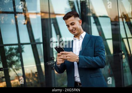 A businessman, wearing a blue suit and white shirt, stands outside of a modern glass office building. He is smiling as he uses his smartphone. Stock Photo