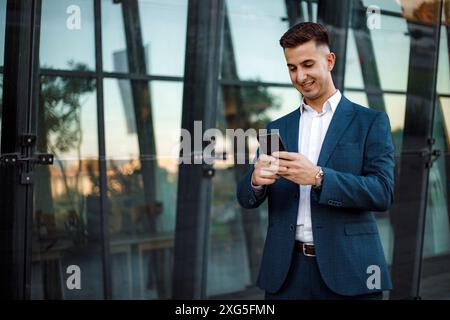 A businessman in a blue suit stands outside a modern building, checking his smartphone. The sun is shining and the sky is clear. Stock Photo