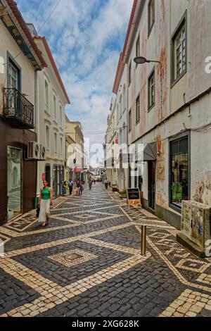 People walk along a narrow cobbled street lined with historic buildings and cafes under a clear sky, Ponta Delgada, Sao Miguel Island, Azores Stock Photo