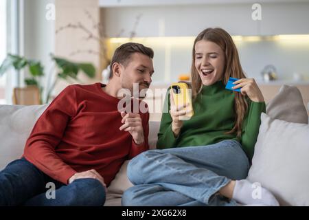 Happy married couple, spouses on couch making online purchases via internet using smartphone. Stock Photo