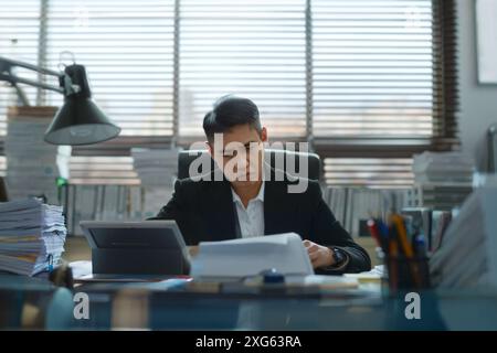 Asian man Concentrated office worker in business suite at working desk littered with paper and stack of documents and laptop in overloaded with work c Stock Photo