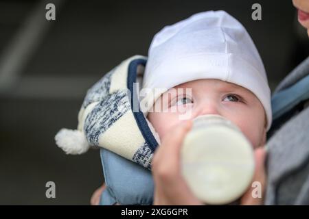 Close-up of a baby drinking from a bottle while being held by the mother. The baby is warmly dressed in a knitted hat and jacket Stock Photo