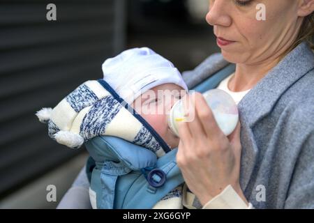 Close-up of a baby being fed from a bottle by the mother, dressed in a gray coat. The baby is warmly dressed in a knitted hat and jacket Stock Photo