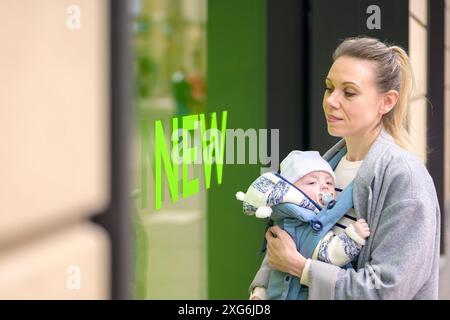 A mother holds her baby in a blue carrier in front of a store window with the word 'NEW' in green letters. The baby, wearing a knitted hat and jacket, Stock Photo