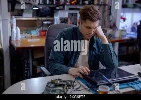 caucasian professional repairman sit thinking how to repair fault laptop, sit behind table at workshop. Repairs and diagnostics breakdowns of laptop i Stock Photo